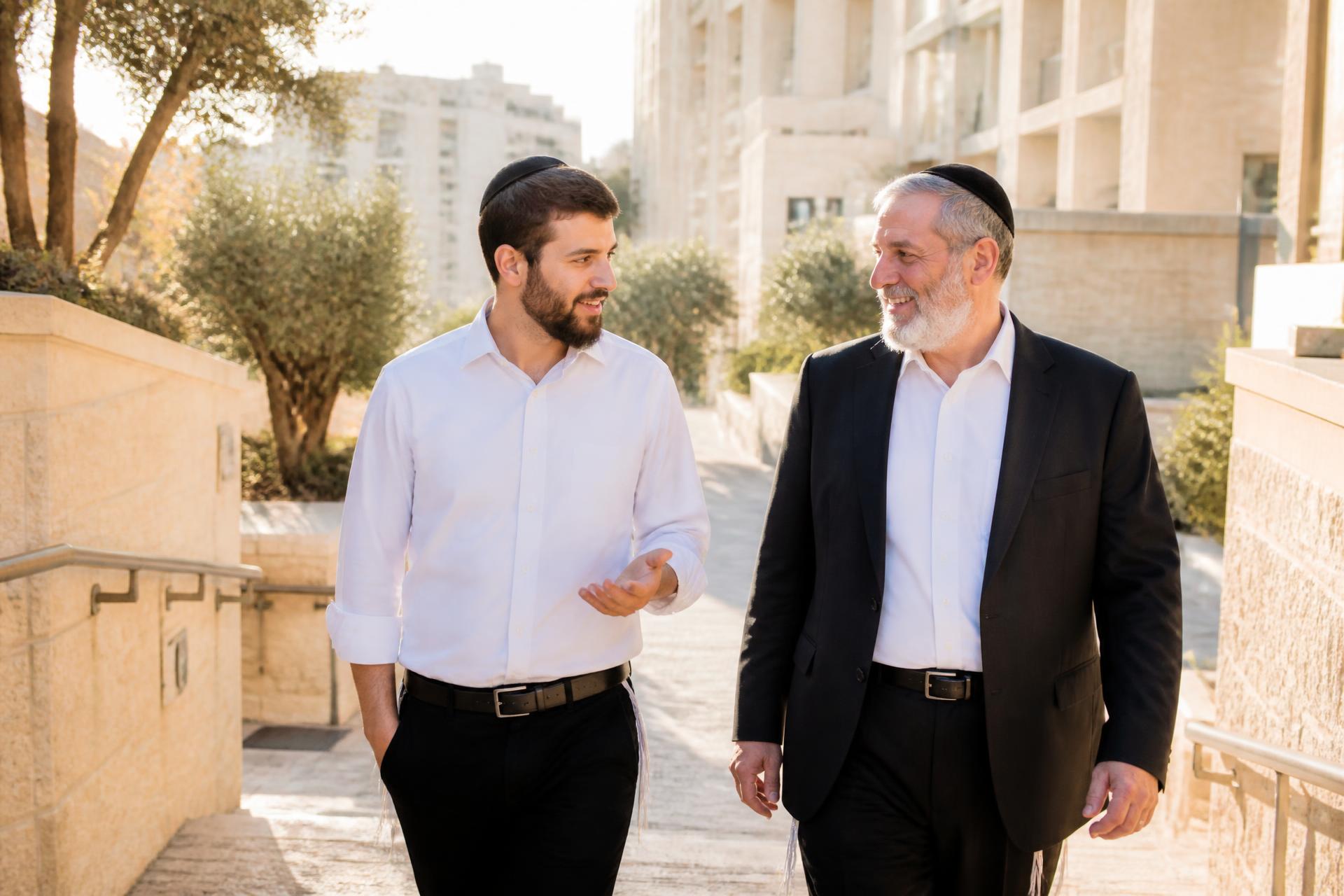 A young Orthodox Jewish man walking and talking warmly with his father on a sunlit Jerusalem street.